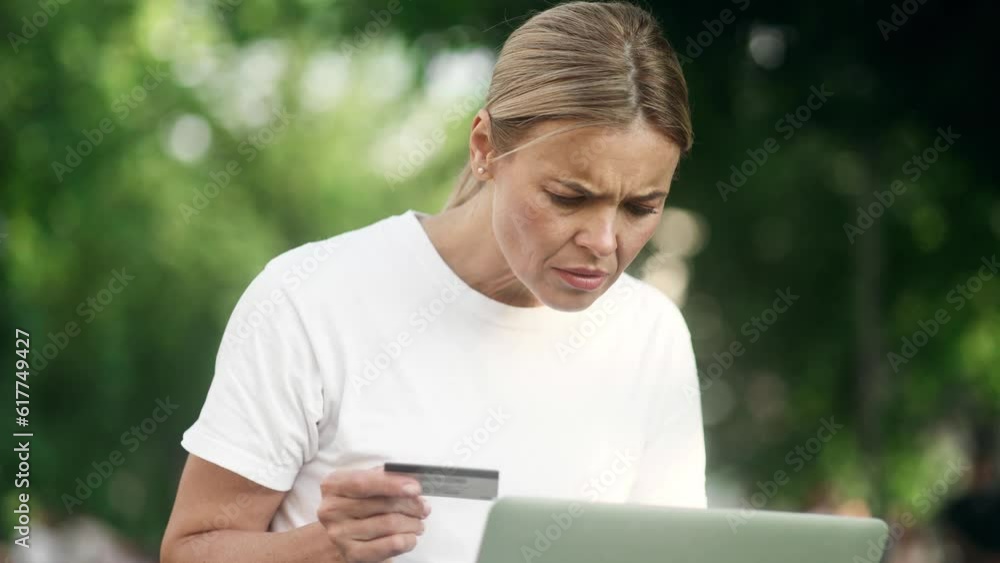 Confused blond woman with credit card thoughtfully pondering on camera and shrugs while has problem with online order payment error or blocked bank account doing shopping at internet on laptop at park Confused blond woman with credit card thoughtfully pondering on camera and shrugs while has problem with online order payment error or blocked bank account doing shopping at internet on laptop at park