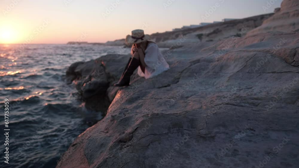 Huge rocks at Mediterranean sea with blurred woman in white dress and straw hat sitting at background admiring golden sunset. Unrecognizable Caucasian female tourist in twilight dusk on vacations Huge rocks at Mediterranean sea with blurred woman in white dress and straw hat sitting at background admiring golden sunset. Unrecognizable Caucasian female tourist in twilight dusk on vacations