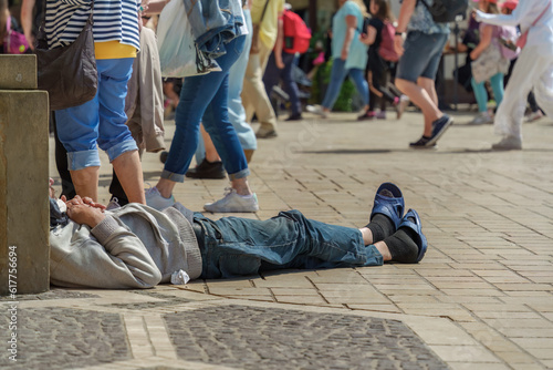 Authentic tramp sleeping on a stone-paved street on a sunny summer day against the background of a large group of passers-by and tourists, people walk past a needy man lying on the city street