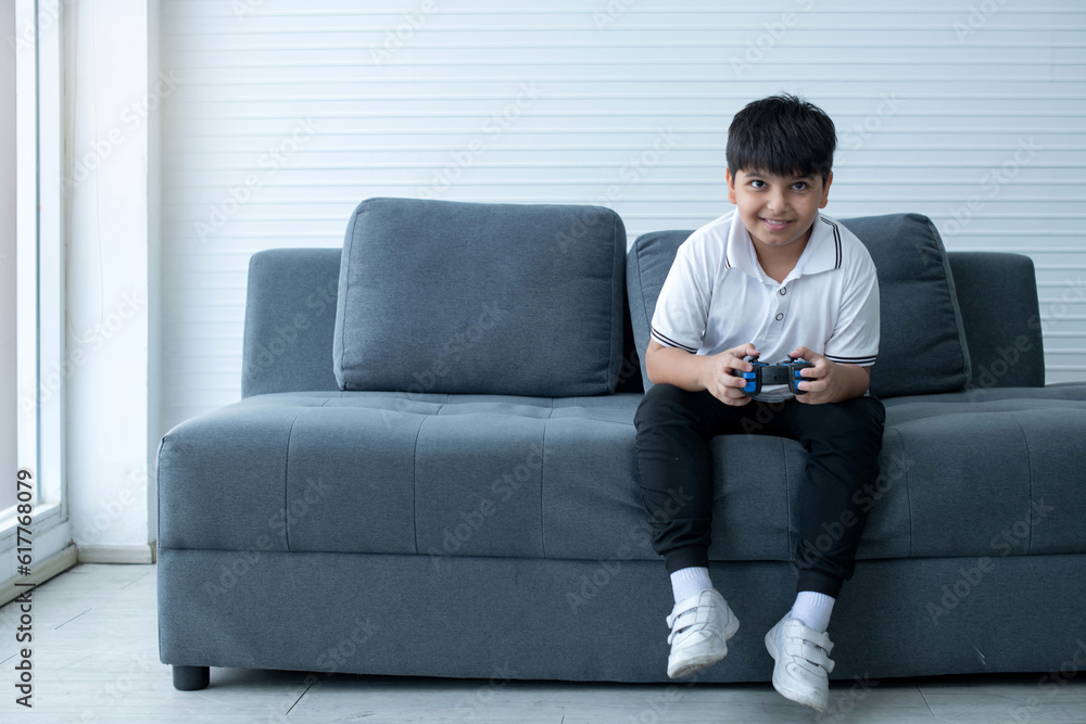 Concentrated Indian boy excited playing video game, sitting on couch and holding controller having fun in gamer emotion, kid gaming addiction concept