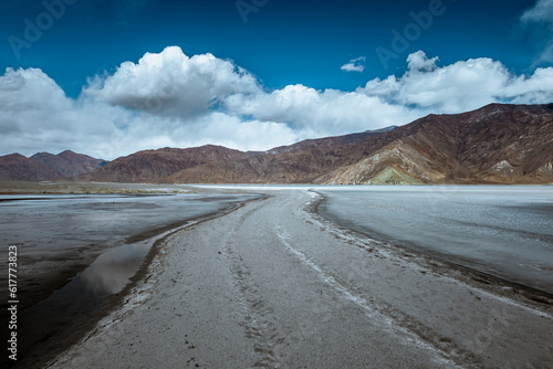 A walkway amidst the fully frozen Pangong Lake, inviting you to explore the captivating natural landscape on a sunny day, under a clear blue sky adorned with fluffy white clouds © PrabhjitSingh