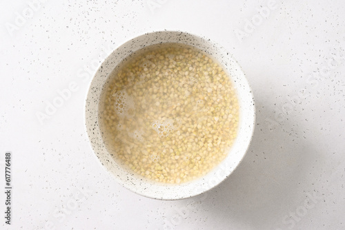 Making vegan milk of green buckwheat. Soaking process grain with water in white bowl on white background. Close up. Copy space.