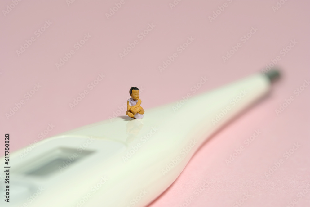 Miniature tiny people toy figure photography. A girl toddler baby seating above thermometer while whimpering on pink background Miniature tiny people toy figure photography. A girl toddler baby seating above thermometer while whimpering on pink background