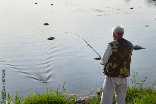 Old man fishing. Senior gray haired fisherman throws a spinning from shoreside at sunset, twisting a coil. Positive elderly male angling at lake, rotating reel rod. Retired fisher enjoying his weekend