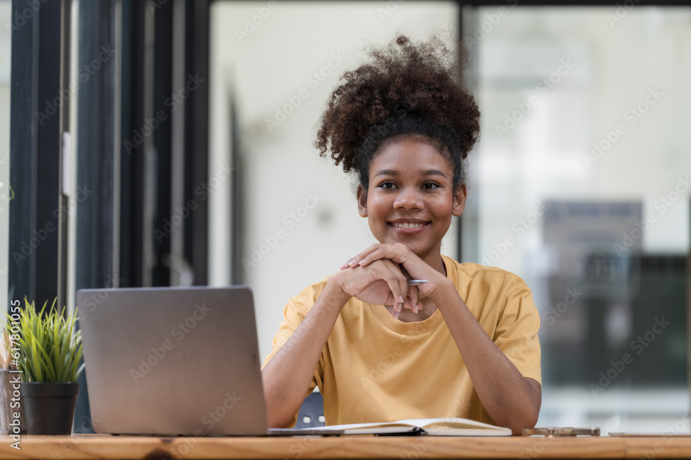 A young African American stylish woman, freelancer, student, or real estate agent, sitting at her desk at home office, looking at the camera and smiling pleasantly. A young African American stylish woman, freelancer, student, or real estate agent, sitting at her desk at home office, looking at the camera and smiling pleasantly.