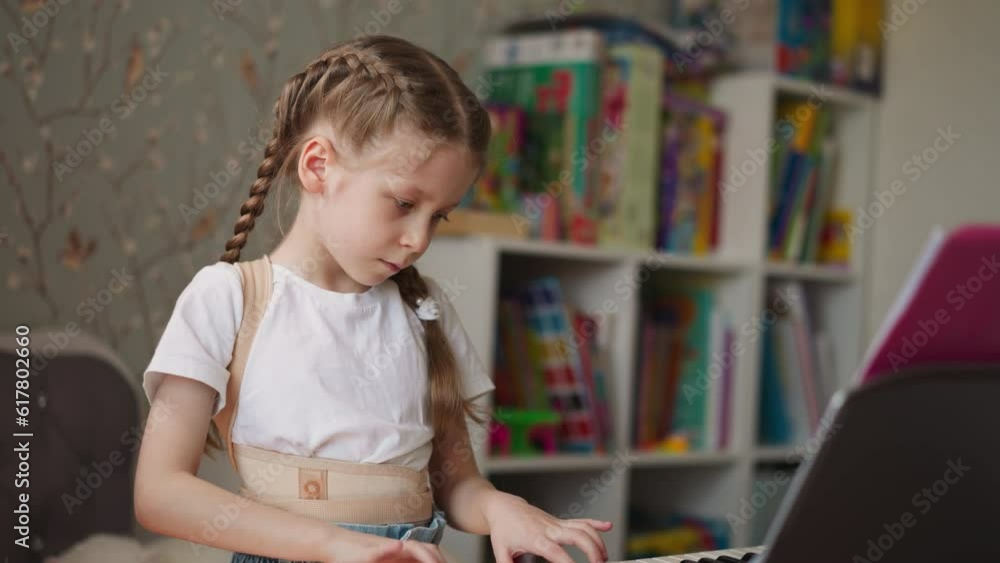 Cute little girl with spinal injury plays piano making careful smooth movements. Graceful child with long braids practices music in cozy nursery closeup Cute little girl with spinal injury plays piano making careful smooth movements. Graceful child with long braids practices music in cozy nursery closeup