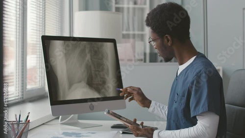 Waist up of young African American male doctor in blue scrubs looking at computer monitor while analyzing patient spine x-ray photo