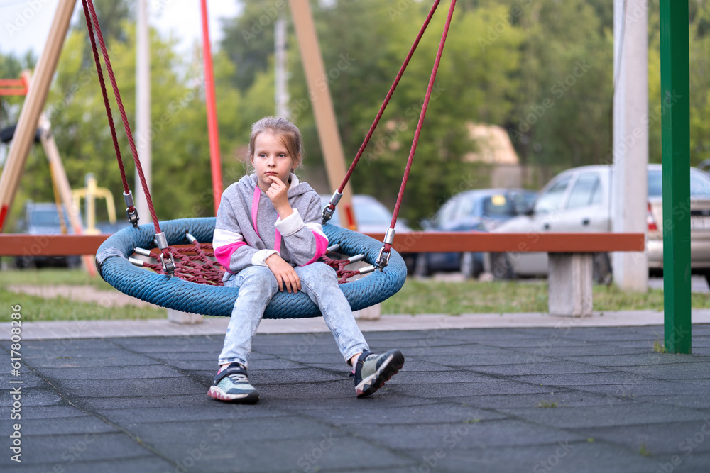A teenage sad girl with long hair pulled back in a ponytail, wearing jeans, a hoodie and sneakers is sitting on a round swing on a sunny summer day.The concept of loneliness, problems of adolescence. A teenage sad girl with long hair pulled back in a ponytail, wearing jeans, a hoodie and sneakers is sitting on a round swing on a sunny summer day.The concept of loneliness, problems of adolescence.