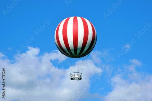 red and white gas filled huge balloon against blue sky in Budapest, city park. white clouds. round metal observation deck with tourists. summer scene. leisure and outdoors. abstract low angle view.