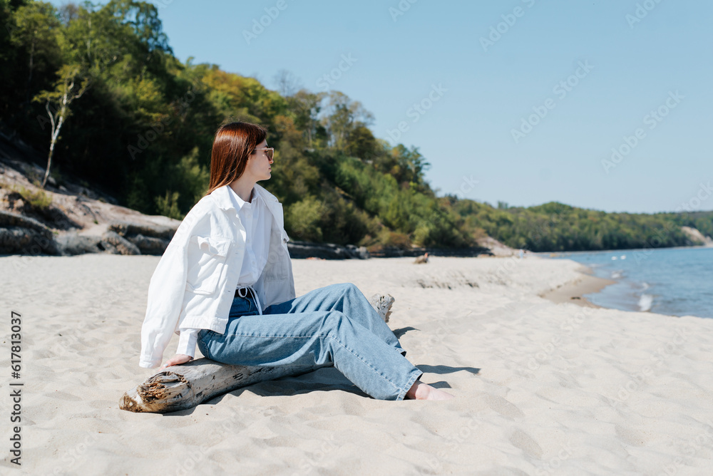 Lonely beautiful young woman in a jacket and jeans sitting on a log on shore. Side view of a barefoot woman resting on seashore, mood Lonely beautiful young woman in a jacket and jeans sitting on a log on shore. Side view of a barefoot woman resting on seashore, mood