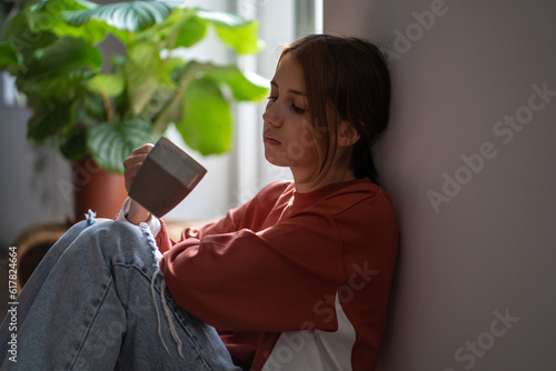 Frustrated sad teenage girl sitting on floor with cup devastated thinking about trouble, broken heart. Young woman have bad mood due to hormones in period. Depressive thoughts from problems at school. photo