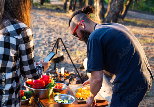 Stylish man with a beard and black glasses cuts fresh vegetables peppers and tomatoes to prepare a salad in nature. Vacation with friends in nature. Tourism as a hobby. Evening sunset.