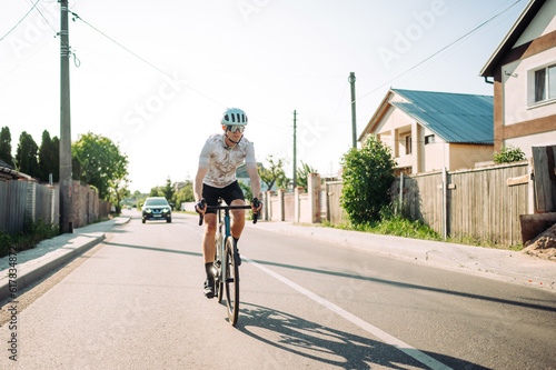 A professional cyclist rides a bicycle on a road through a town with houses and a car in the background.