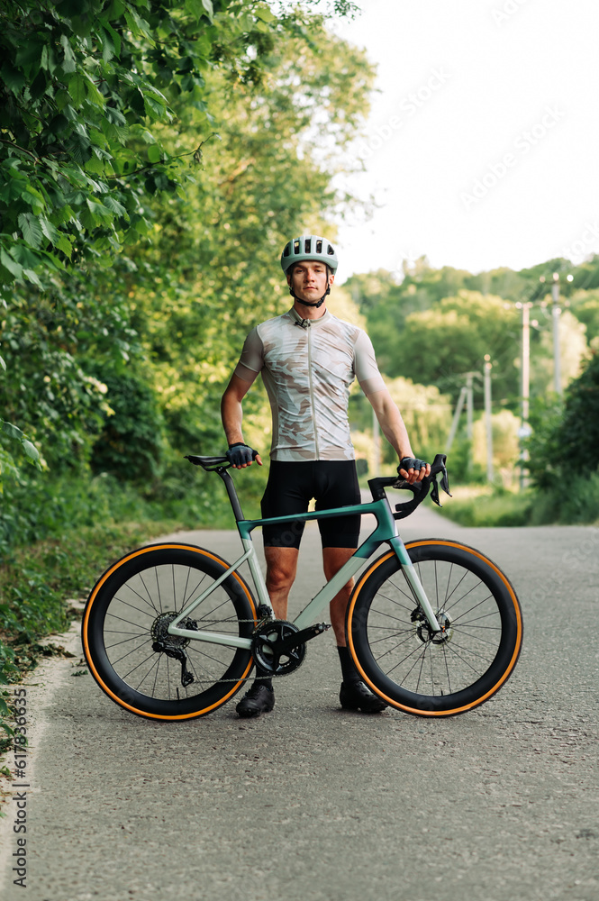 Obraz premium Photo of a sporty male cyclist in gear posing for the camera with a professional road bike outside the city on a road and forest background.