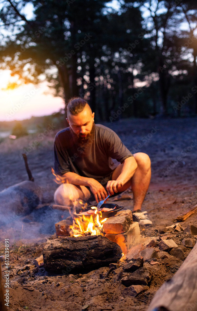 A young man with a beard prepares a dish of bacon with scrambled eggs on a fire. Cooking and outdoor recreation in summer. A young man with a beard prepares a dish of bacon with scrambled eggs on a fire. Cooking and outdoor recreation in summer.
