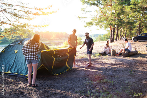 A company of friends guys and girls set up a tent on the sandy bank of the river in summer. Hiking, outdoor recreation in the forest. Sunny day.