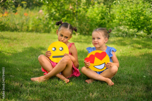One shy girl holds a sad tearful emoticon, the second a smile face with hearts instead of eyes