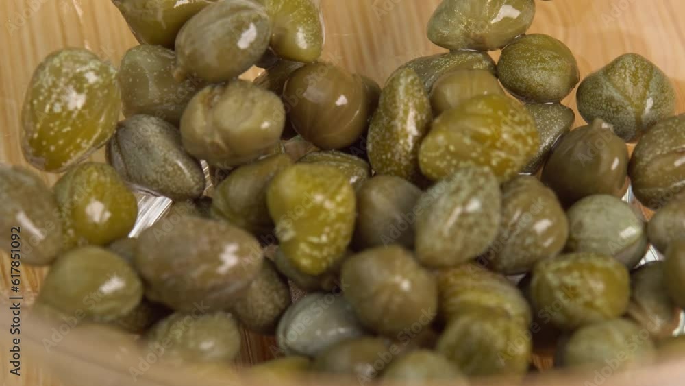Pickled marinated capers pouring in glass bowl on wooden background. Ingredients for making spicy sauce, tartare or salad