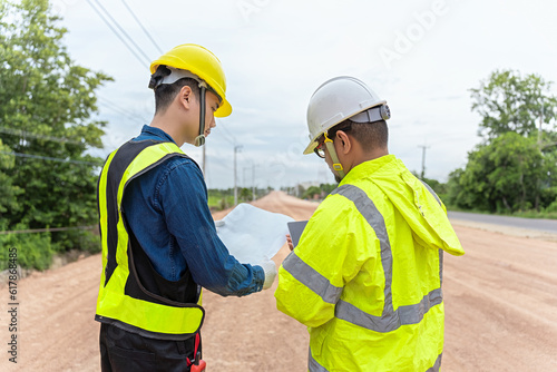 Asian engineer holding tablet and foreman holding road construction blueprints, on construction site to supervise road construction In the background is a road under construction and machinery working