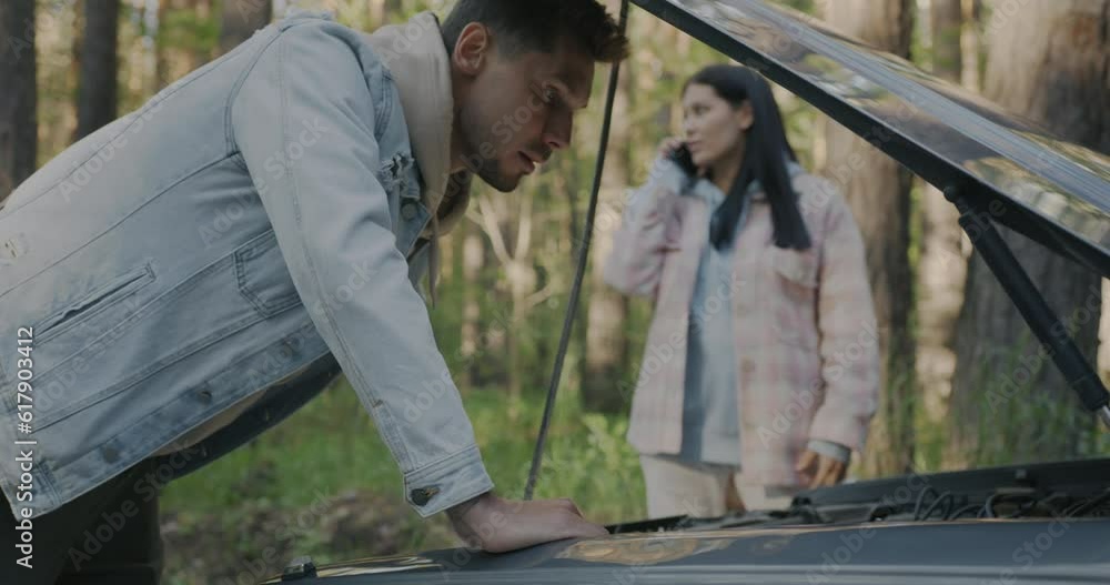 Man repairing broken car while woman calling for emergency service asking for help in forest. Vehicle trouble and road trip concept. Man repairing broken car while woman calling for emergency service asking for help in forest. Vehicle trouble and road trip concept.