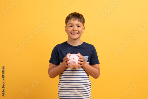 Smiling boy holding piggy bank for saving, isolatd on yellow