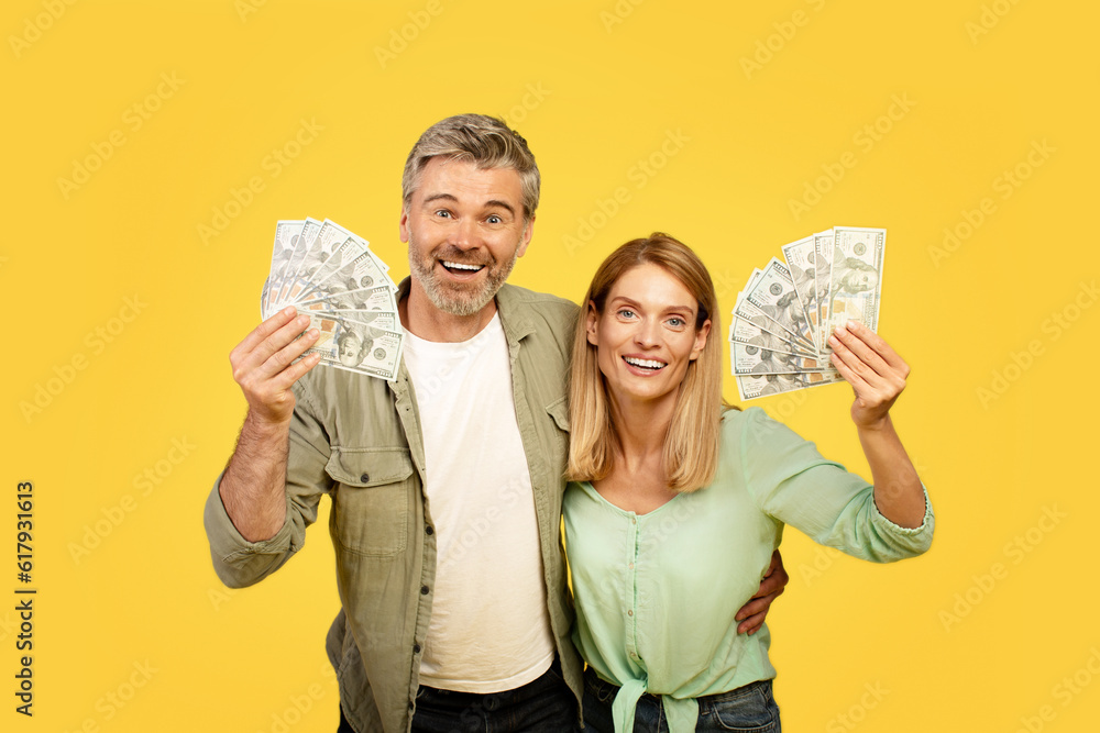 Overjoyed european middle aged couple holding fans of dollar cash, smiling at camera on yellow studio background Overjoyed european middle aged couple holding fans of dollar cash, smiling at camera on yellow studio background