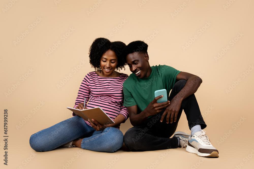 Positive young african american husband with phone, pregnant wife with big belly making notes in notebook Positive young african american husband with phone, pregnant wife with big belly making notes in notebook