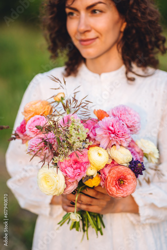 Portrait of attractive young beautiful woman with curly hair in a long white lace dress holding bouquet of bright colourful ranunculi flowers. Summer evening, sunset golden hour summertime inspiration