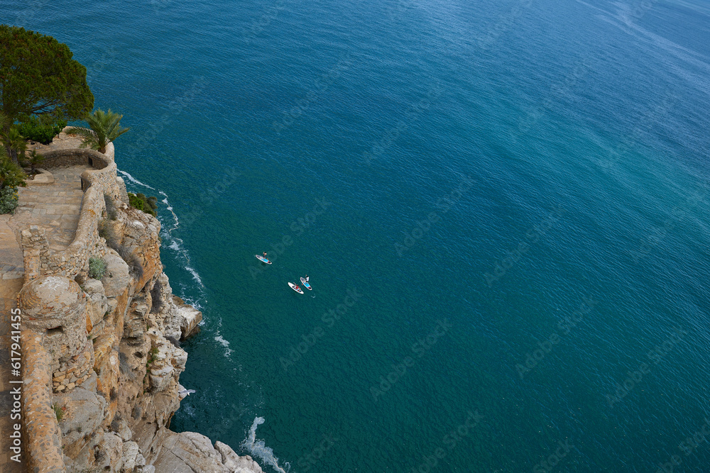 3 people are rowing on SUP boards along castle wall on a rocky cliff with giant stones.