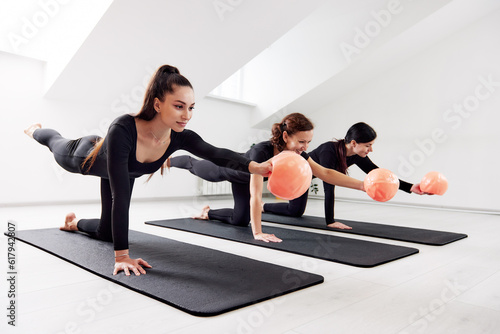 Beautiful women are doing Pilates in a bright studio. Three female athletes in black tracksuits doing exercises with orange balls on yoga mats. Group Pilates classes. Healthy lifestyle.