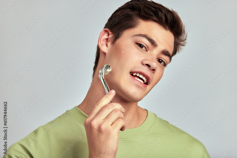 A young handsome guy with brown hair doing self-care routine using a face massage roller in a studio. Caucasian man caring for the skin to maintain his youthful and healthy appearance. Men's skincare. A young handsome guy with brown hair doing self-care routine using a face massage roller in a studio. Caucasian man caring for the skin to maintain his youthful and healthy appearance. Men's skincare.