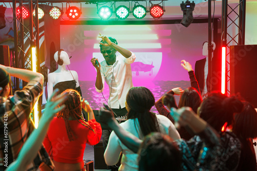 Crowd dancing and raising hands while musician performing on stage in club. People moving on dancefloor and having fun while enjoying live electronic music concert in nightclub