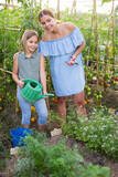 Young woman with clipper and her daughter with watering pot standing in kitchen garden and smiling.