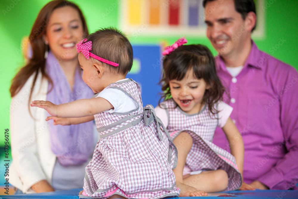 Obraz premium Beautiful ten months baby girl playing with her sister and their parents on background.. Early stimulation for toddlers and children concept.