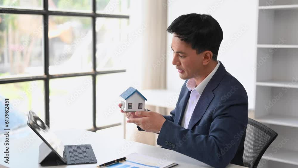 Young real estate agent worker working with laptop and tablet at table in office and small house beside it.. Young real estate agent worker working with laptop and tablet at table in office and small house beside it..