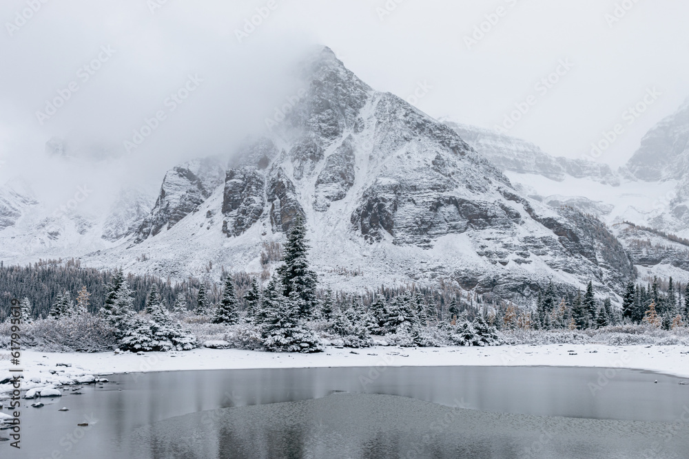 Fototapeta premium vue sur une montagne en forme de triangle recouverte de neige lors d'une journée grise avec les nuages bas et un lac en avant plan