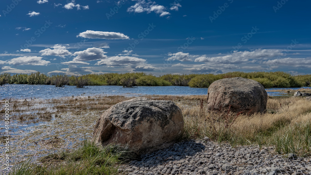 Boulders on the shore of Redonda Bay. Dry grass around. The soil is reinforced with a metal mesh. The blue water of the lake glistens in the sun. Green bushes, azure sky with clouds. Argentina. 
