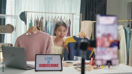 Young Asia lady seller look at phone camera live stream use laptop answer follower comment introduce sweater new collection new arrival on her chanel at studio. Woman fashion online shop concept.