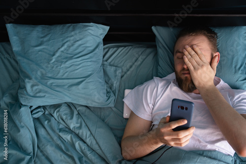 Confused man looking on mobile phone lying in bed at home in the morning. Oversleeping concept. photo