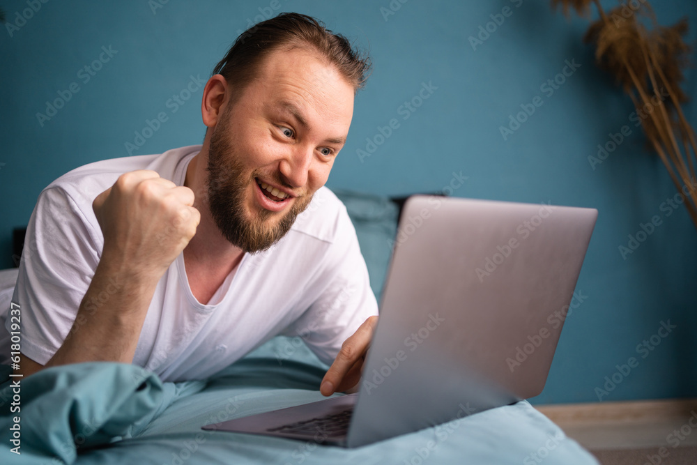Portrait of happy man spending his weekend laying in bed, watching soccer play live broadcast on his laptop and celebrating money win from bets on his favourite team. Portrait of happy man spending his weekend laying in bed, watching soccer play live broadcast on his laptop and celebrating money win from bets on his favourite team.