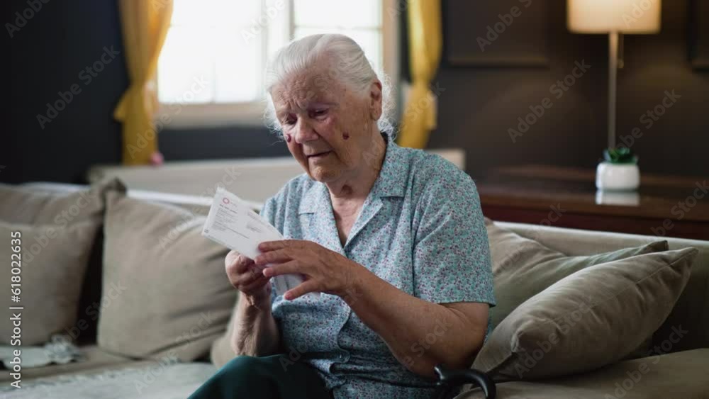 Frustrated shocked old 80s retired woman looking at large utility bills sitting on the sofa in the room at home, problem in the household budget and financial expenses Frustrated shocked old 80s retired woman looking at large utility bills sitting on the sofa in the room at home, problem in the household budget and financial expenses