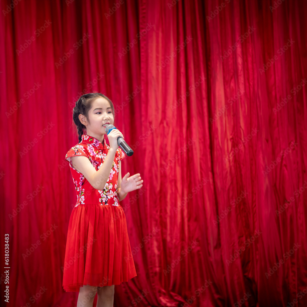 The Asian kid girl sing a song on stage at her school activity day, dress in Qipao style, red curtain background The Asian kid girl sing a song on stage at her school activity day, dress in Qipao style, red curtain background