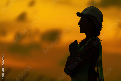 Half body silhouette of female engineers smiling and holding laptop stand near the industrial plant The twilight sky resembles a sunset, glowing with golden light, wearing a helmet and vest.