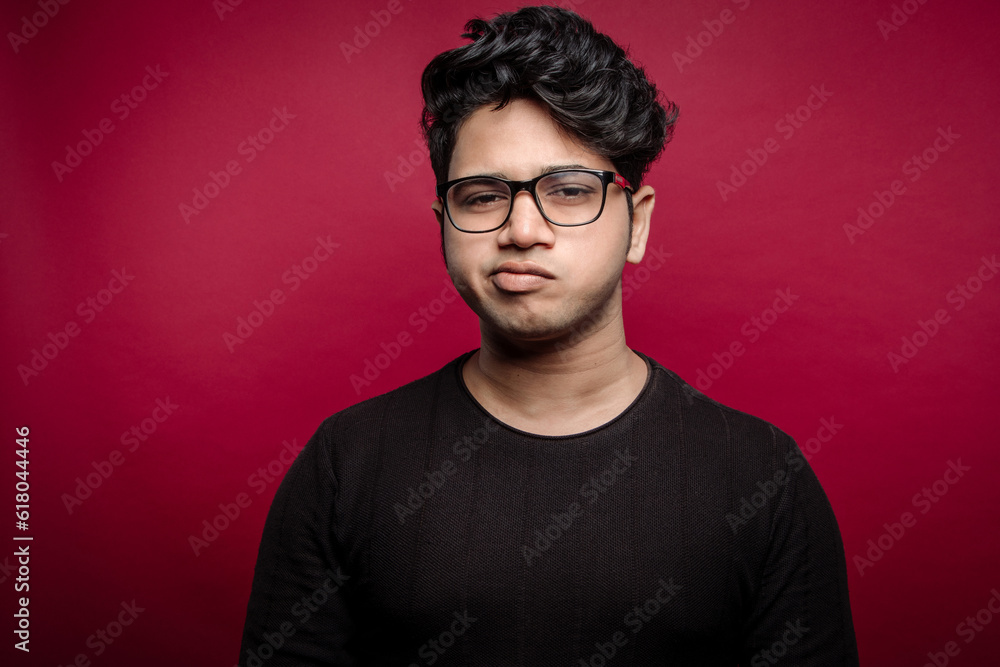 A young man displays clear disapproval on his face, with furrowed brows and a tight-lipped expression, showcasing his discontent and disagreement Isolated On A Red Studio Background.