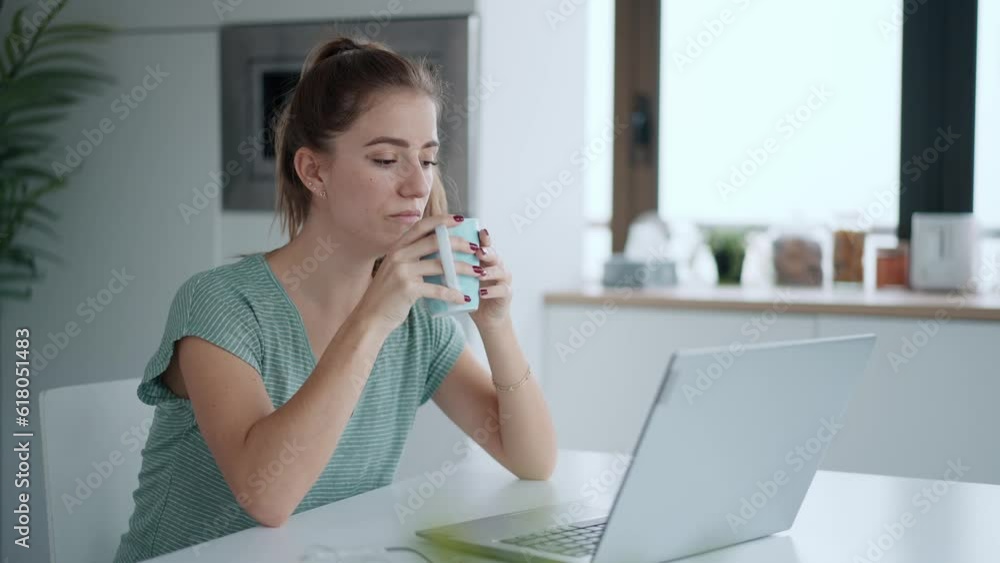 Video of pretty young woman working with laptop while drinking cup of coffee in the kitchen at home. Video of pretty young woman working with laptop while drinking cup of coffee in the kitchen at home.
