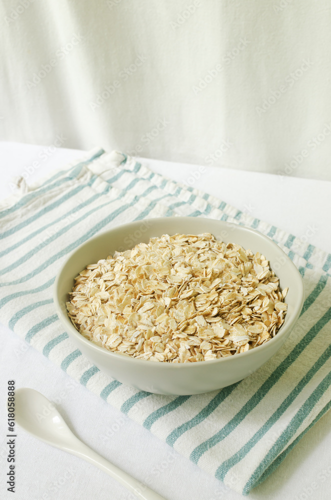 Raw oatmeal in a gray bowl on a white tablecloth with a kitchen napkin and a teaspoon. Concept of healthy eating. Vegetarian and vegan food. Vertical orientation. Selective focus. Raw oatmeal in a gray bowl on a white tablecloth with a kitchen napkin and a teaspoon. Concept of healthy eating. Vegetarian and vegan food. Vertical orientation. Selective focus.