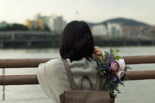 A girl stands at the side of a river with a bouquet of flowers on her hand. A girl looks at the city from a deck at the bank of a river with blurry buildings in the background © Emanuele