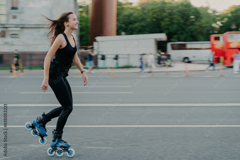 Energetic slim young smiling woman rollerblades on city road enjoys spending free time actively moves quickly has dark hair floating on wind leads healthy lifestyle. Active weekends. Outdoor photo Energetic slim young smiling woman rollerblades on city road enjoys spending free time actively moves quickly has dark hair floating on wind leads healthy lifestyle. Active weekends. Outdoor photo