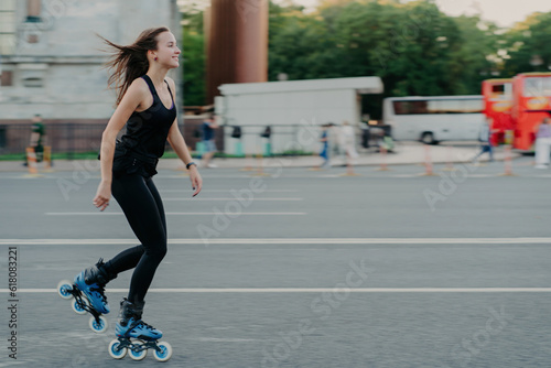 Energetic slim young smiling woman rollerblades on city road enjoys spending free time actively moves quickly has dark hair floating on wind leads healthy lifestyle. Active weekends. Outdoor photo