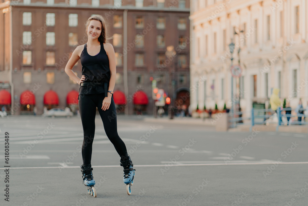 Sport and recreation concept. Horizontal shot of active woman practices rollerblading poses on blades keeps hand on waist blurred city background takes break. Rolleskating after work. Healthy life